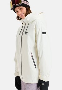 Witte waterdichte jas met capuchon, zwarte ritsen en logo-detail. Gecombineerd met zwarte handschoenen met een slanke, gladde textuur.