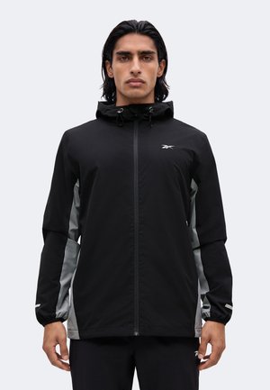 Mann trägt eine schwarz-graue Kapuzenjacke mit Reißverschluss und elastischen Bündchen, steht frontal vor einem einfarbigen hellen Hintergrund.