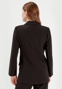 Black tailored blazer with a structured collar, long sleeves, and a single back vent. Smooth texture and fitted design emphasize a sleek silhouette.