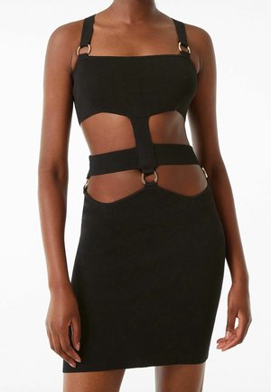 Femme portant une robe noire à découpes avec des bretelles épaisses et des détails en anneaux métalliques, exposant le ventre et le haut des cuisses sur un fond blanc.