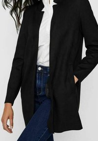 Cappotto in suede nero con collo alto, maniche lunghe, tasche laterali e taglio dritto. Indossato sopra una camicia bianca e jeans in denim.
