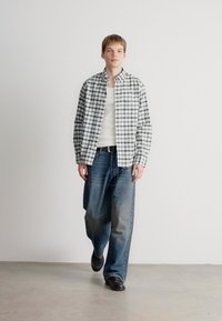 Rutet langermet skjorte i grønn og hvit, brukt over et hvitt tanktop. Løse blå denimjeans, svarte sko, minimalistisk bakgrunn.