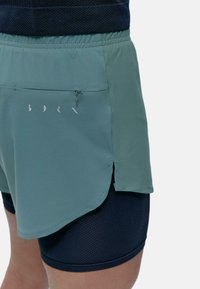 Grüne Sportshorts mit hohem Bund, Reißverschlüssen und seitlichen Schlitzen, die über schwarzen, strukturierten Kompressionsshorts getragen werden.