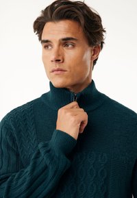 Teksturowany sweter w kolorze teal z wysokim kołnierzem i zamkiem. Posiada wzory w plecionkę oraz kontrastujące gładkie sekcje. Długie rękawy z prążkowanymi mankietami.