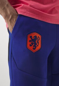 Blaue Jogginghosen mit einem auffälligen orangefarbenen Löwenwappen und "KNVB"-Schriftzug, aus glattem Stoff und mit seitlichen Reißverschlusstaschen. Teilweise sichtbares pinkes Shirt.