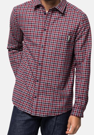 Mand iført en rød, blå og hvid ternet langærmet skjorte med sorte knapper og mørkeblå jeans, stående med den ene hånd i lommen.