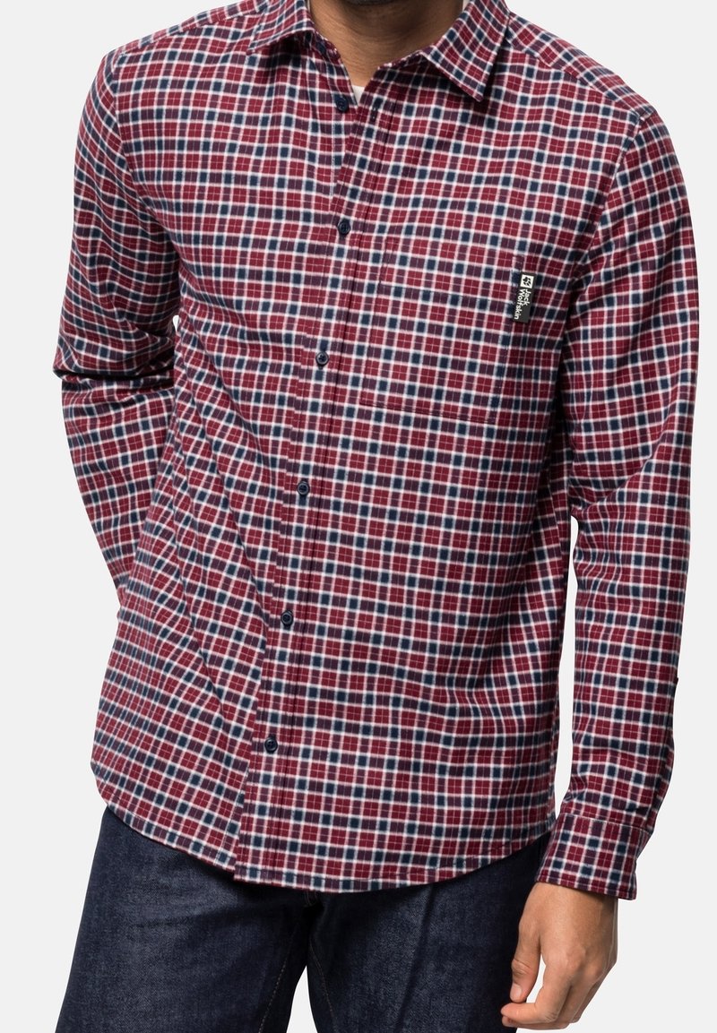 Homme portant une chemise à carreaux rouge, bleue et blanche à manches longues avec des boutons noirs et un jean bleu foncé, debout avec une main dans la poche.