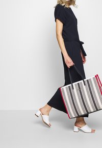Gestreifte Tragetasche mit schwarzen und weißen vertikalen Streifen, roten Kanten und schwarzen Trägern. Getragen mit einem marineblauen Outfit und weißen High Heels.