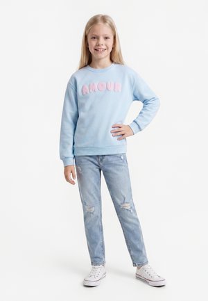Blondes Mädchen trägt ein hellblaues Sweatshirt mit rosa "AMOUR" Buchstaben, zerrissene hellblaue Jeans und weiße Sneakers, steht mit einer Hand in der Hüfte.