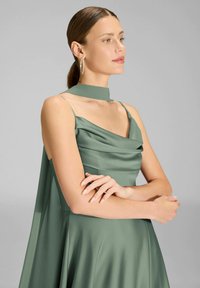 Vestito in raso verde con corpetto drappeggiato, collo alto e spalline sottili. Presenta una gonna fluente e una texture liscia. Orecchini circolari visibili.