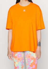 Oranžové bavlněné tričko s volným střihem, krátkými rukávy a malým bílým logem Adidas na hrudi. Hladká textura, minimalistický design.