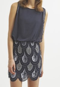 Haut sans manches bleu marine avec une silhouette fluide, associé à une jupe courte avec un design en sequins ondulés argentés et bleu marine foncé.