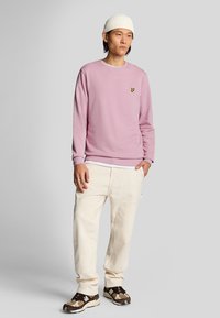 Sudadera rosa claro con cuello redondo y logo amarillo bordado, combinada con pantalones cargo en color blanco sucio y zapatillas marrones.