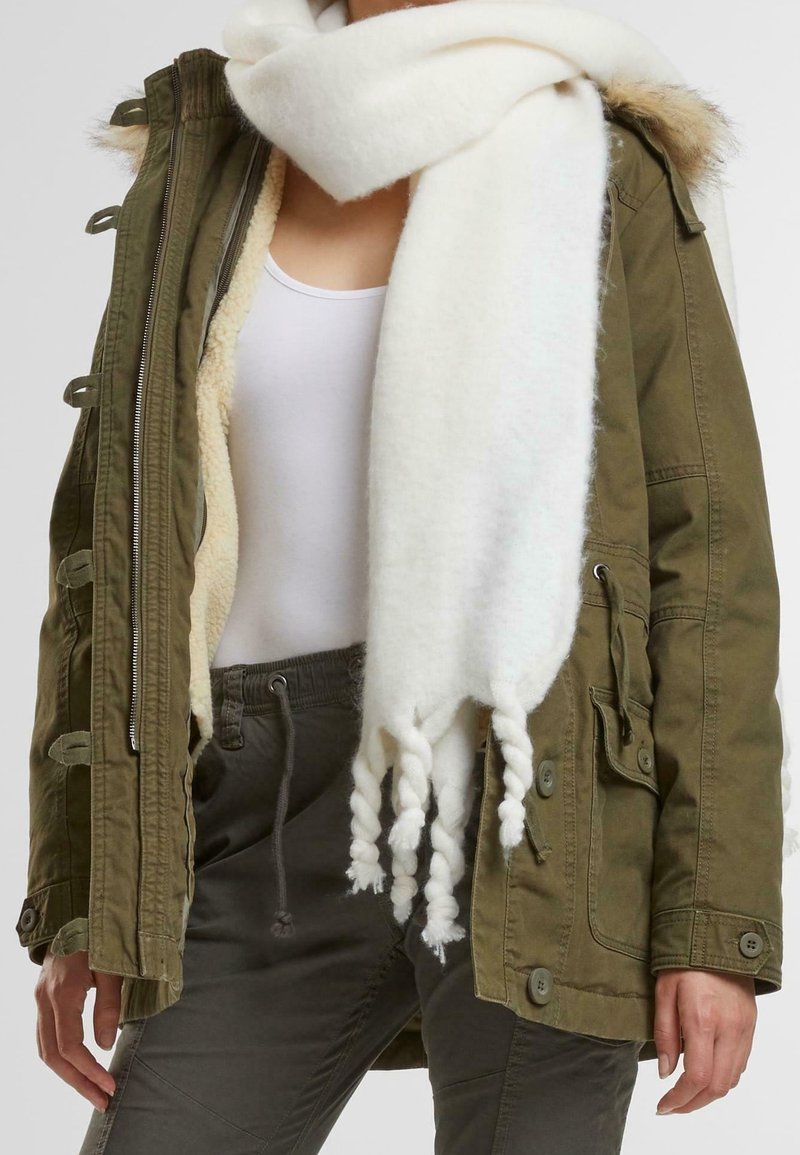 Olive green parka jacket with shearling lining, zip closure, and pockets. Soft white scarf with twisted fringe edges. Gray drawstring trousers.