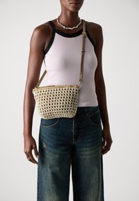 Woven bag in cream with a textured pattern, featuring a golden zipper, brown adjustable strap, and a round shape. Paired with a light tank top and jeans.