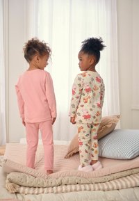 Kaksi lasta pyjamassa seisoo sängyllä. Toinen on pukeutunut vaaleanpunaiseen pitkähihaiseen paitaansa ja housuihin; toinen puolestaan käyttää kuviollista kermaista paitaa ja housuja, joissa on sarjakuvahahmoja.