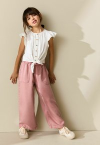 Balts īsroku krekls ar pogām un sasienamu priekšpusi, kombinēts ar vaļīgiem rozā džinsa biksēm. Modelis valkā gaišas krāsas sporta apavus. Neitrāla fona.