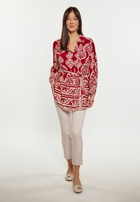 Rød og creme mønstret bluse med lange ærmer, krave og bælte i taljen, parret med lys beige slim bukser og flade sko.