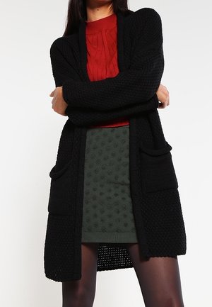 Personne portant un long cardigan noir texturé sur un pull rouge torsadé et une mini-jupe verte à motifs, debout les bras croisés.
