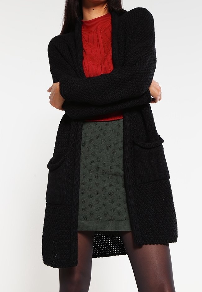 Personne portant un long cardigan noir texturé sur un pull rouge torsadé et une mini-jupe verte à motifs, debout les bras croisés.