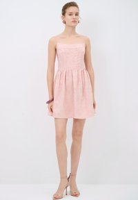Vestido rosa claro con un corpiño ajustado y una falda acampanada, que presenta una tela texturizada y tirantes finos, exhibido en un modelo en un entorno neutro.