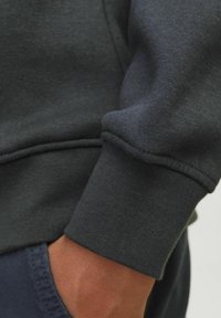 Polsino di felpa grigio scuro che mostra la trama a costine e dettagli di cucitura, con una finitura liscia e una mano consumata appoggiata su un tessuto più scuro.