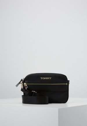 Zwarte rechthoekige crossbodytas met gouden rits en "TOMMY"-logo, geplaatst op een witte ondergrond tegen een lichtgrijze achtergrond.