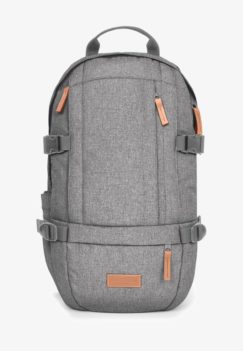 Sac à dos en tissu gris avec un design structuré, des bretelles réglables, des accents en cuir, des tirettes de fermeture éclair et des poches latérales.