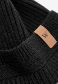 Maglione nero a costine con collo alto e un'etichetta in pelle con la lettera "W". Il tessuto appare morbido e strutturato.