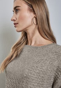 Beige stickad tröja med ribbat och texturerat mönster, med en vid halsringning. Modellen har långt hår och bär silverfärgade hoopörhängen.