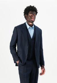 Junger Mann mit Vitiligo, der einen marineblauen Anzug und ein hellblaues Hemd trägt, steht vor einem weißen Hintergrund mit einer Hand in der Tasche.