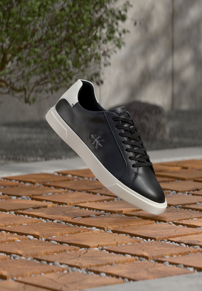 Ασπρόμαυρο αθλητικό παπούτσι Calvin Klein με κορδόνια, κρεμασμένο πάνω από ξύλινο και πέτρινο μοτίβο σε εξωτερικό χώρο.