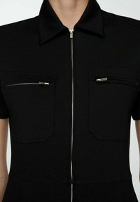 Sort jumpsuit med korte ærmer. Udstyret med en lynlås foran, to brystlommer med lynlåse og en krave. Glat, tætsiddende stof.