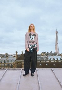 Cămașă cu dungi roșii și albe cu mâneci lungi, având un grafic cu Mickey Mouse, asortată cu pantaloni negri largi și pantofi negri. În fundal, orizontul orașului Paris.