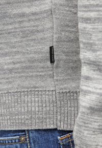 Pull gris tricoté avec un ourlet côtelé et une étiquette portant le nom de la marque "Jack & Jones". La texture est lisse avec des variations de couleur subtiles.
