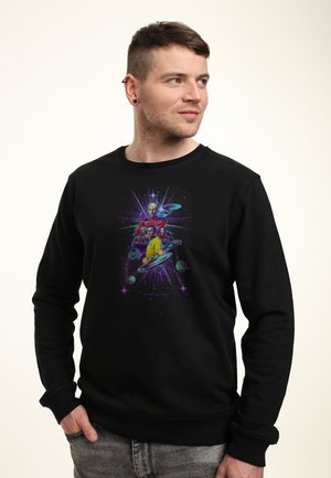 Uomo con capelli corti e piercing alle orecchie indossa una felpa nera con personaggi di fantascienza colorati e astronavi, guardando di lato.