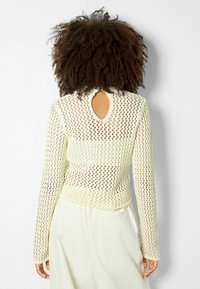 Stickad tröja i en ljus citronfärg, med ett mönster av diamantöppningar, långa ärmar och en cirkelrund nyckelhålsutskärning bak.