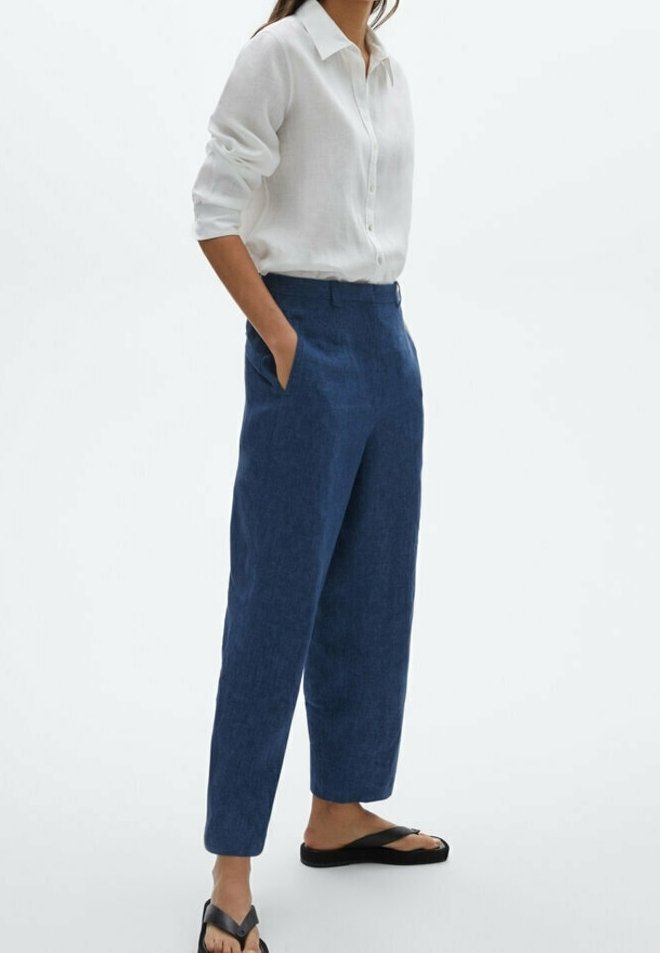 Femme portant une chemise blanche boutonnée aux manches retroussées, un pantalon bleu taille haute et des tongs noires, debout les mains dans les poches.