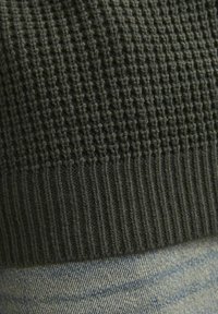Tessuto in maglia verde scuro e testurizzato con fondo a coste, abbinato a denim azzurro chiaro, leggermente usurato, che mostra una cucitura visibile.