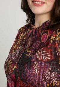 Blusa floreale in un intenso bordeaux scuro con vivaci motivi gialli, rosa e verdi. Presenta un collo arricciato con chiusura a fiocco e un tessuto leggero.