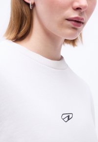 Κοντινό πλάνο ενός ατόμου που φοράει λευκό φούτερ με στρογγυλή λαιμόκοψη, με ένα μικρό μαύρο γεωμετρικό λογότυπο στο στήθος και ένα ασημένιο κρίκο σκουλαρίκι.