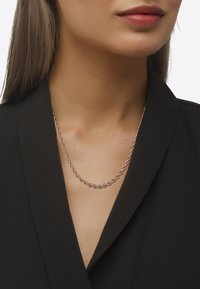 Collana a catena metallica torsionata di colore oro rosa, appoggiata su un collo nudo sopra un blazer nero, che mette in risalto il suo design elegante.