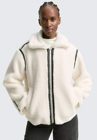 Jachetă albă din fleece cu închidere cu fermoar, dungi negre de accent pe mâneci și guler deschis. Textură moale și croială supradimensionată.