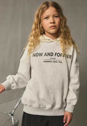 Blond kind met golvend haar draagt een lichtgrijze hoodie met tekst over vriendelijkheid en liefde, staand naast een doorzichtige stoel tegen een effen achtergrond.
