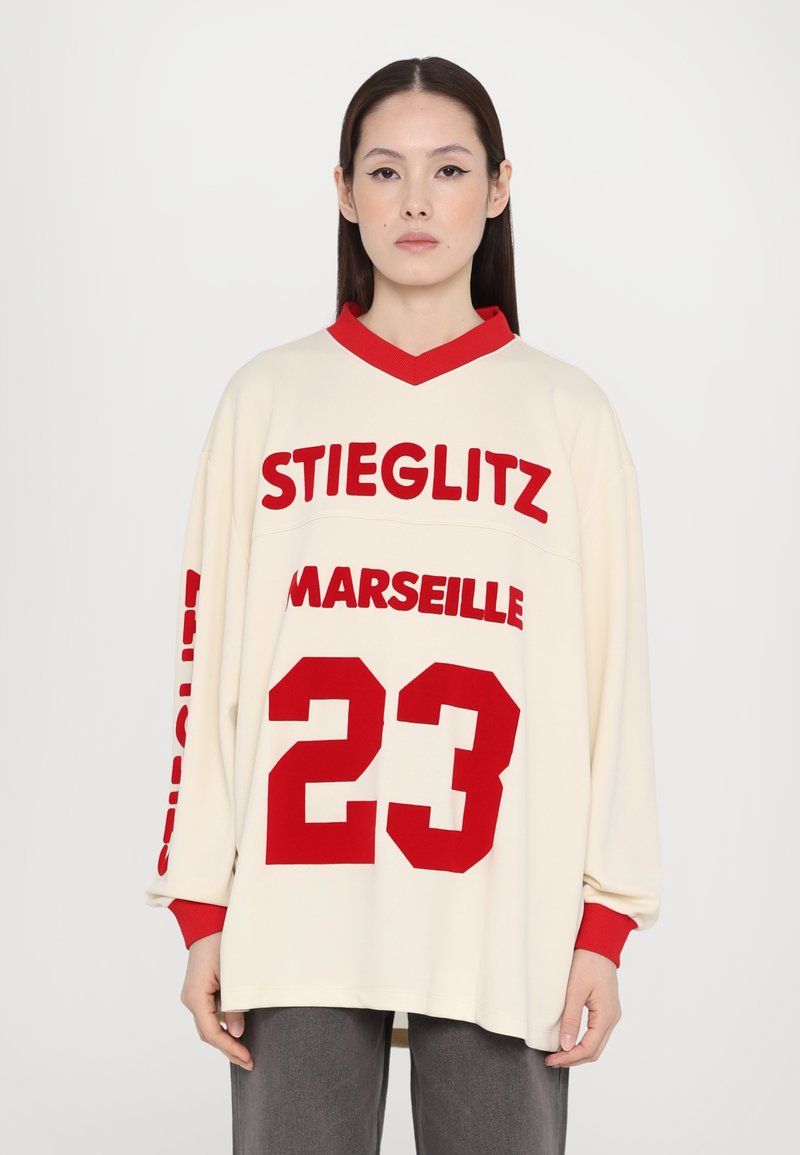 Nadmerná krémová jersey s červeným textom: "STIEGLITZ" a "MARSEILLE," číslo "23," s červeným rebrovaným golierom a manžetami. Mäkko textúrovaná, voľnočasový dizajn.