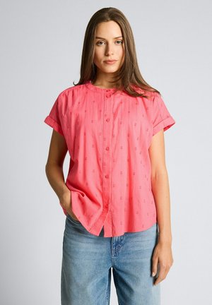 Kobieta z długimi brązowymi włosami, ubrana w koralową koszulę na guziki z krótkim rękawem i niebieskie dżinsy, stoi z jedną ręką w kieszeni.