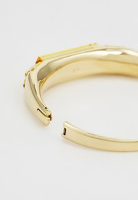 Bracelet doré avec un design courbé, présentant une surface texturée et des accents angulaires en jaune. Comprend un détail de fermeture à agrafe.