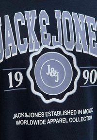 Tums zilā krāsā, izgatavots no kokvilnas, ar lieliem baltajiem un gaiši purpura burtiem, uz kuriem rakstīts "JACK & JONES", apaļš logo un dibināšanas gads "1990".