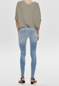 Femme portant un jean skinny bleu clair, un haut ample en maille taupe et des sandales à talons noires, debout de dos sur un fond gris uni.
