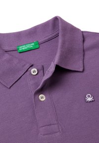 Lila Poloshirt aus strukturiertem Stoff, mit einem Kragen, zwei weißen Knöpfen und einem kleinen gestickten Logo auf der linken Seite. Das Etikett zeigt den Markennamen.
